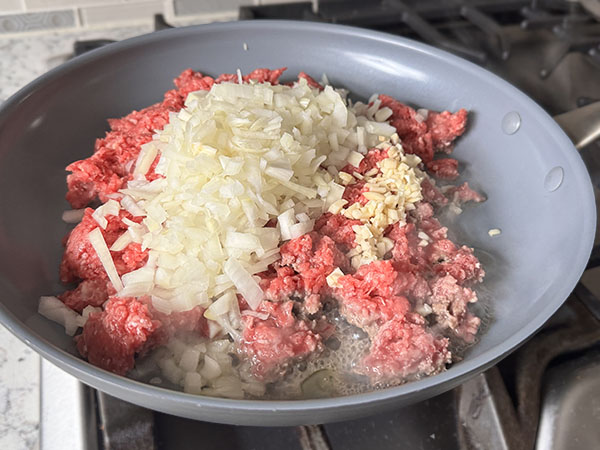 Raw ground beef in a skillet with chopped onions and minced garlic.