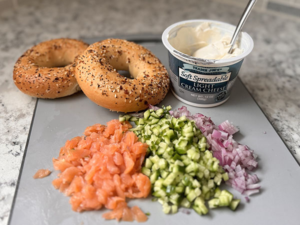 Finely chopped onion, cucumber and lox, along with bagels and cream cheese spread on a cutting board.