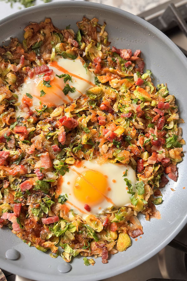 Breakfast hash skillet with over easy eggs, crispy potatoes, Brussels sprouts and onions; topped with bacon bits, hot sauce and fresh chopped parsley.