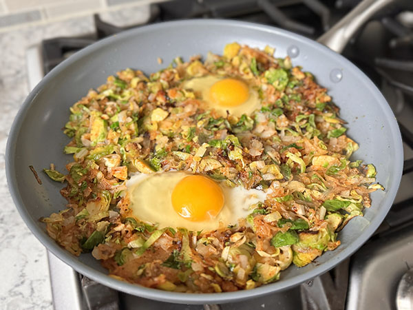 Eggs cracked in the prepared wells in the Brussels sprouts and potato hash cooking in a skillet.