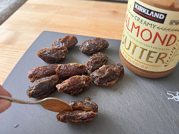 Dates being filled with almond butter using a spoon, with a container of creamy almond butter next to them.