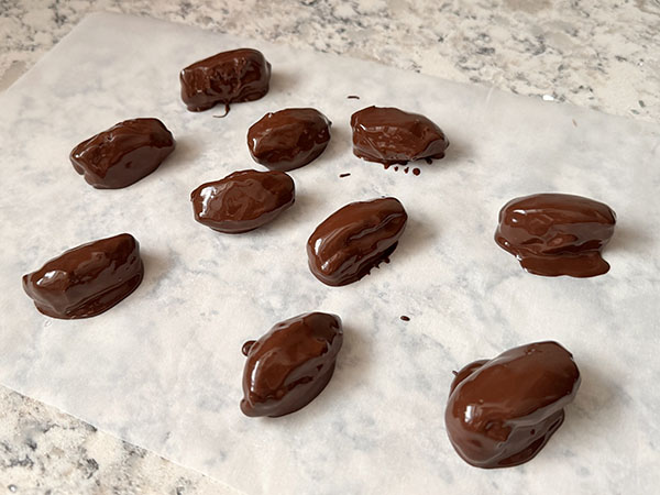 Chocolate covered, almond butter stuffed dates on a piece of parchment paper.
