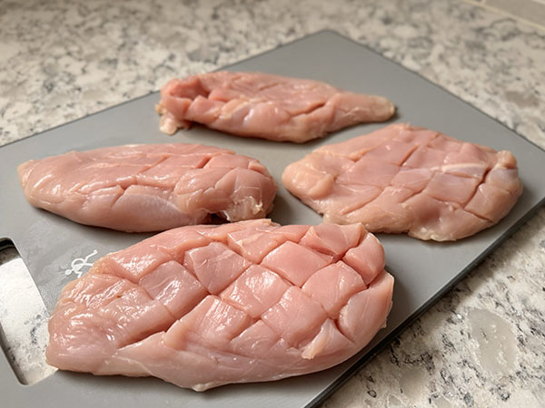 Scored raw chicken breasts on a cutting board.