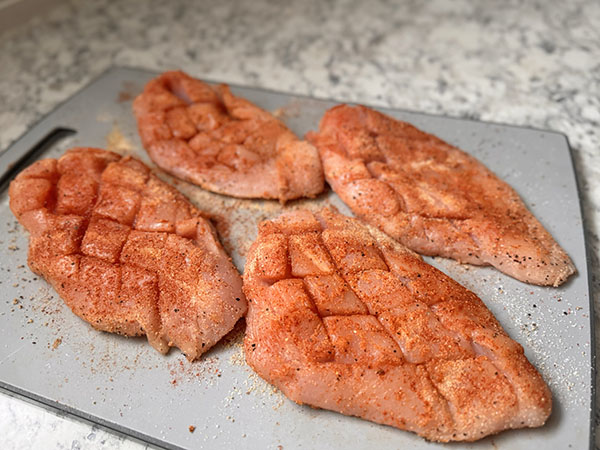 Raw chicken breasts, scored and seasoned, sitting on a cutting board.