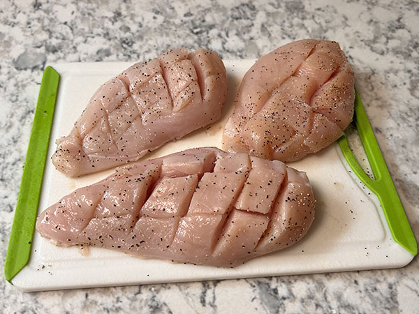 Raw boneless skinless chicken breasts with crosshatch pattern cuts on a cutting board.
